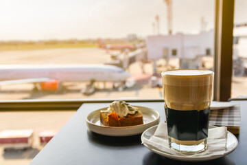 Hot coffee with breakfast at airport lounge, cup of hot cappuccino coffee in a glass cup on the table in the airport lobby, coffee and cake with plane background.