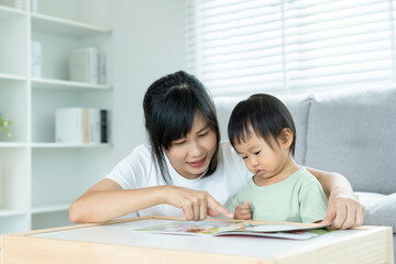 Obraz na płótnie Canvas Happy Asian mother relax and read book with baby time together at home. parent sit on sofa with daughter and reading a story. learn development, childcare, laughing, education, storytelling, practice.