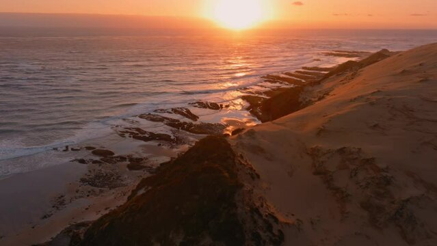 Aerial: The Hokianga Harbour and ocean surf crashing on the shoreline and giant sand dune at sunset. Opononi, Northland, New Zealand.