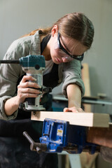 Fototapeta na wymiar Female Carpenter Wearing Protective Safety Glasses and Using Electric Work on a Wood. Artist or Furniture Designer Working on a Product Idea in a Workshop.