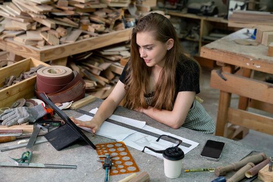 A young woman is training to be a carpenter in the workshop. She works with a laptop computer in a wood workshop. female carpenter contact customers by smartphone. SME orders, Start-up and small