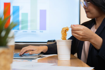 Happy Asian businesswoman has to eat noodles while working on laptop at office.