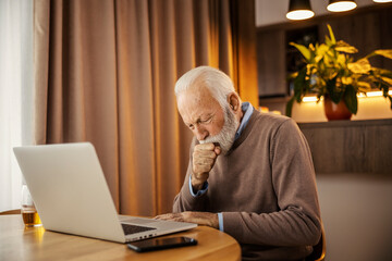 A sick senior man is sitting at home with a laptop and coughing.