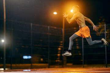 An urban shirtless muscular runner is jumping and running on cold rainy weather at night.