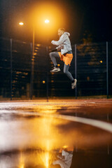 Fototapeta na wymiar A fit urban sportsman is jumping high on sports ground on rainy cold weather at night.