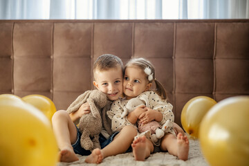 Cute brother and sister are sitting with their toys and hugging surrounded by balloons.