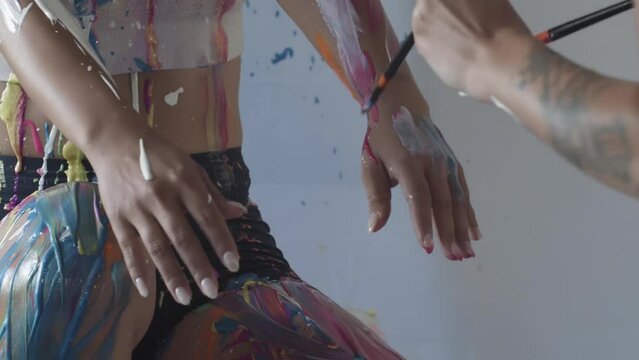 A tattooed woman is slowly applying colorful body paint onto her model friend