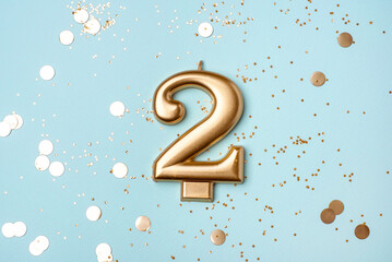 Gold candle in the form of number two on blue background with confetti.