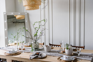 Happy easter dinner with decor at home. Minimalist composition in living room with copy space, hanging lamp, candle, easter eggs, vase with twig, branch, leaves. Interior design of easter dining room