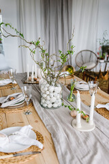 Setting table for Easter dinner with candle, ceramic plates with easter eggs in nest, bunny made of linen napkin. Holidays celebration concept. Decor with vase with twig, branch, leave at home. Set up