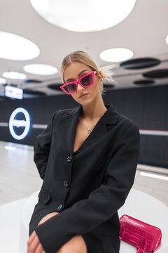 Beautiful sexy woman in fashion pink sunglasses in a stylish black jacket sits and poses indoors. Modern urban chic girl