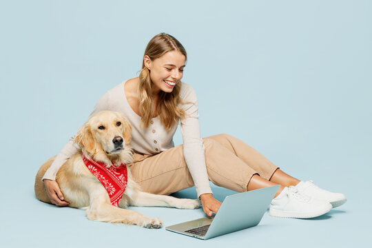 Full body happy fun young owner IT woman wear casual clothes sit near her best friend retriever dog work using laptop pc computer isolated on plain blue background studio. Take care about pet concept.