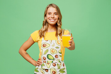 Young happy fun housewife housekeeper chef cook baker woman wear apron yellow t-shirt hold in hand give glass drink orange juice isolated on plain pastel green background studio. Cooking food concept.