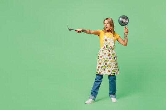 Full body mad sad young housewife housekeeper chef cook baker woman wear apron yellow t-shirt hold frying pan spatula posing pov fight isolated on plain pastel green background. Cooking food concept.