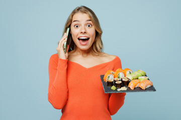 Young surprised woman wears orange casual clothes talk speak on mobile cell phone hold eat raw fresh sushi roll served on black plate Japanese food isolated on plain blue background studio portrait.