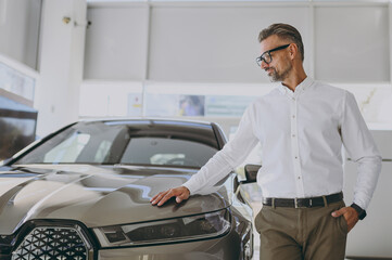 Adult rich happy man customer male buyer client wearing shirt put hand on hood loot at car choose...
