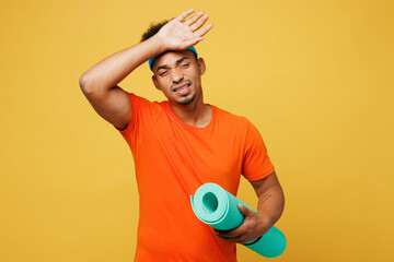 Young tired sad fitness trainer instructor sporty man sportsman wear orange t-shirt put hand on head hold yoga mat train in home gym isolated on plain yellow background. Workout sport fit abs concept.