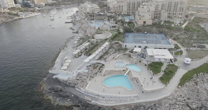 Aerial view of a seaside resort with multiple pools, a tennis court, and moored boats, nestled between a rocky shore and upscale buildings.