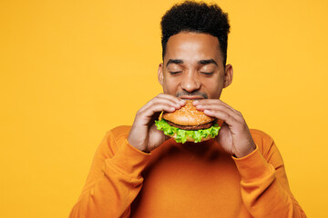 Close up young man wears orange sweatshirt casual clothes holding eating biting burger isolated on...