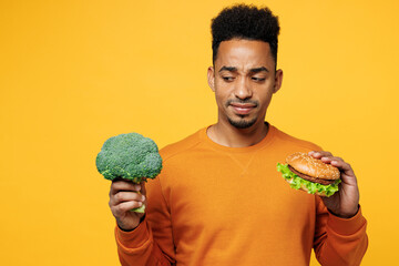 Young confused sad man wear orange sweatshirt casual clothes hold broccoli burger choose what to...