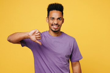 Young upset, displeased man of African American ethnicity wears purple t-shirt casual clothes showing thumb down dislike gesture isolated on plain yellow background studio portrait. Lifestyle concept. - obrazy, fototapety, plakaty
