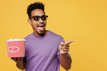 Young surprised fun man of African American ethnicity he wears purple t-shirt casual clothes 3d glasses watch movie film hold bucket of popcorn in cinema isolated on yellow background studio portrait. - obrazy, fototapety, plakaty