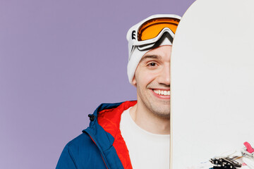 Close up man wear blue windbreaker jacket ski goggles mask hat hold cover half of face with snowboard look camera spend extreme weekend winter season in mountains isolated on plain purple background.