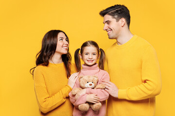 Young happy adorable cute parents mom dad with child kid girl 7-8 years old wear pink knitted sweater casual clothes hug cuddle teddy bear toy isolated on plain yellow background. Family day concept.