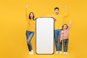 Full body winner happy young parents mom dad child kid girl 7-8 years old wear pink casual clothes big huge blank screen area mobile cell phone isolated on plain yellow background. Family day concept.