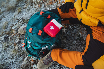 Top view of a red first aid kit, which is lying on a backpack, a tourist in the mountains uses a...