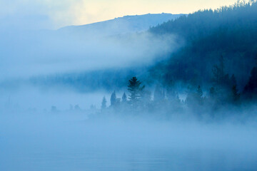Fototapeta premium View over forest with morning fog. Dawn over the foggy lake Yazevoe. Beautiful dreamy view. Lake Yazevoe is located at an altitude of 1685 meters above sea level. Eastern Kazakhstan. Blur effect.