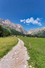 Winding little path along lush meadows with fence and cows leading to a mountain village in Switzerland. Beautiful landscape with farm houses and mountain range on a sunny day in summer.