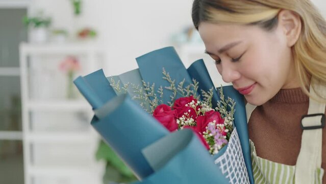 Happy young asian woman florist smelling red rose flowers bouquet at flower shop. Woman florist eyes closed enjoying in smell of bouquet of fresh flowers at flower shop. Small business concept