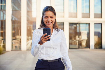Smiling businesswoman walking outside the office using mobile phone texting message