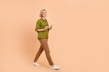 Fototapeta na wymiar Full body photo of lovely girl wear khaki shirt hold cup of latte walk with smartphone to empty space isolated on pastel color background