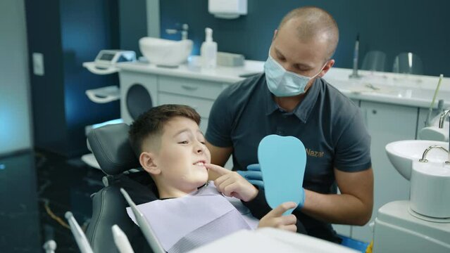 Young male dental expert giving recommmendations to young patient sitting in dentist chair with napkin on chest, holding hand mirror, showing sick tooth. High quality 4k footage