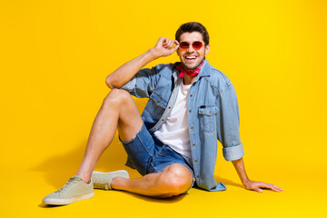 Full body photo of cool optimistic man dressed denim shirt sitting on floor touching stylish glasses isolated on yellow color background