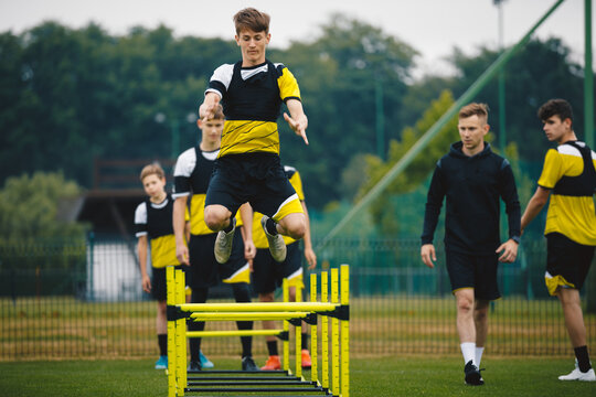 Football camp for youth. Teenage boys in training with a coach at grass pitch. Sports team preparing for the season. Young players jumping over hurdles for strength and coordination training