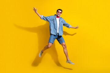 Fototapeta na wymiar Full size photo of good mood guy wear jeans jacket in sunglass jumping hold arms like wings isolated on vibrant yellow color background