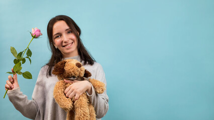 A young smiling woman holding a stuffed toy and a pink rose. Blue cardboard background with copy space for text.