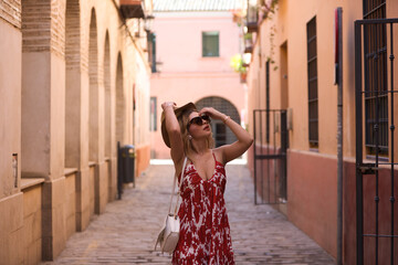 Young and beautiful blonde woman with sunglasses and straw hat on her head walking in the historical centre of seville. The woman is on holiday and enjoys the city. The woman is happy.