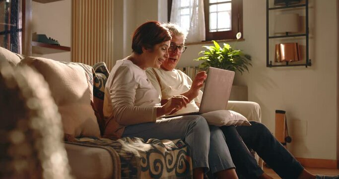 At home, a couple of grandparents make a video call to their relatives and friends using the computer. The elderly couple smiles, blows kisses and is happy to talk to them.
