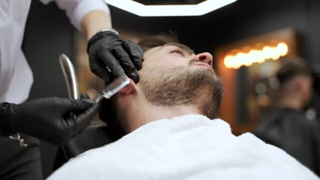 Professional female barber shaves customer beard with razor in upscale barbershop. Man reclines for grooming, stylist focuses on facial hair, luxury setting background. Client relaxes, expert works.