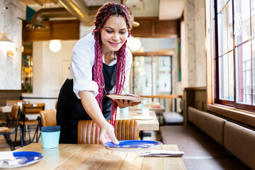 A dark-skinned woman with colorful braided hair wears a waitress's uniform as they set up a table...