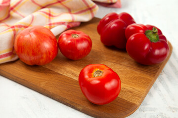 Cutting board with several red tomatoes and bell pepper on white wooden background with red kitchen towel..