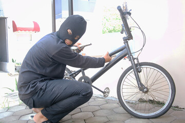 A thief in a balaclava breaks the lock on a bicycle in the street during the day. - obrazy, fototapety, plakaty