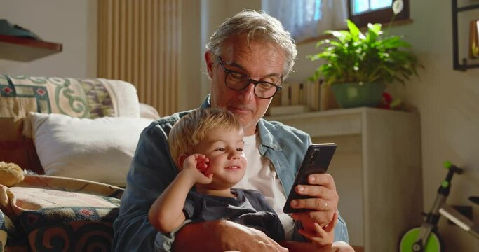 At home, at sunset, the grandfather with his grandson sitting on his lap, looking at the smartphone and laughing together.