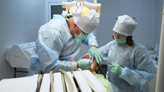 Male dental surgeon, nurse doing medical operation on root canals in operating room. Endodontist and assistant operating patient in modern dental clinic. Oral surgery, installation of dental implants.