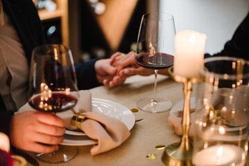Romantic date by candlelight at night. Proposal hand and heart. Hands man and woman hold glasses at home. Toast. Couple in love drinking wine. Cheers. Dinner setup table for couple on Valentine's day. - obrazy, fototapety, plakaty