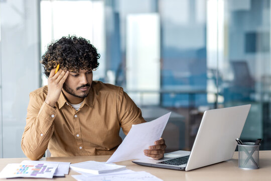Serious thinking and concentrated businessman works with papers contracts reports and invoices, man at workplace for paper work uses laptop, compares data.